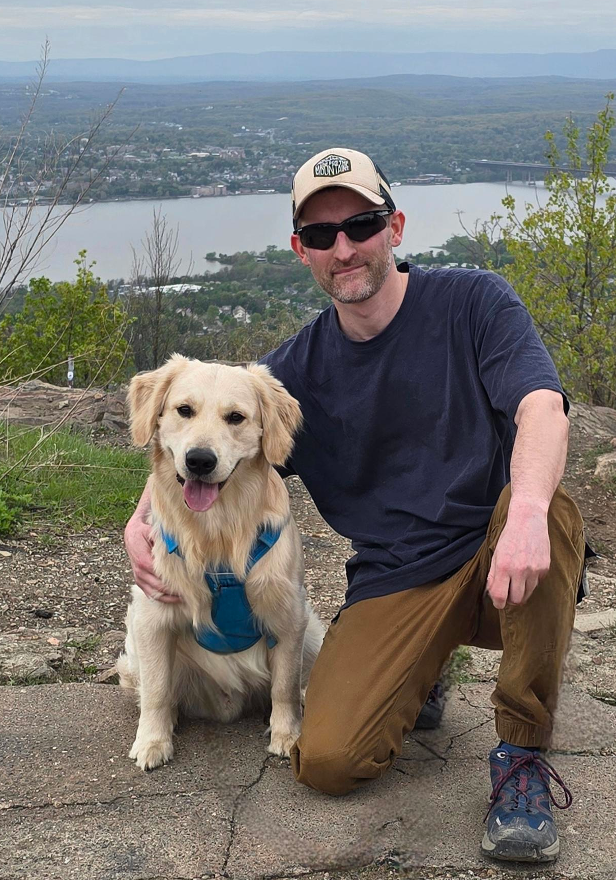 Scott kneeling next to Lincoln, his golden retriever, on a rocky overlook above a river valley